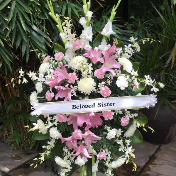 Standing funeral spray with pink lilies and white flowers, labeled "Beloved Sister"