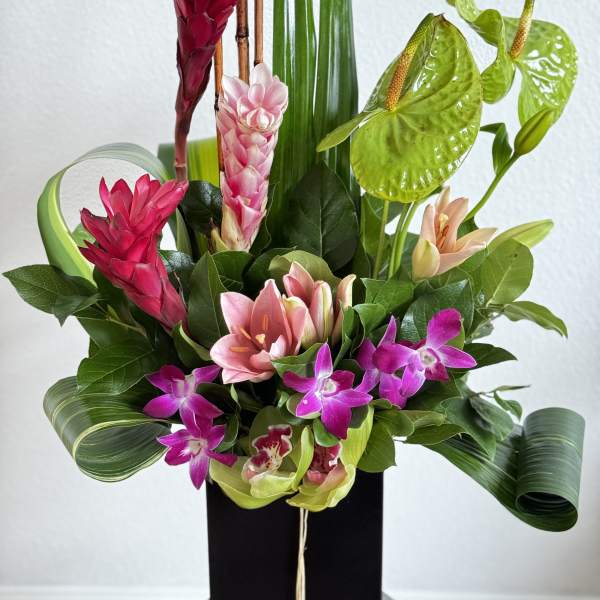 Tall tropical arrangement with pink lilies, orchids, ginger flowers, and sculpted green leaves in a black vase