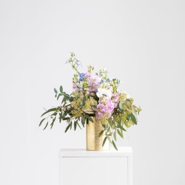 Pastel mixed flower arrangement in a gold cylinder vase on a white pedestal