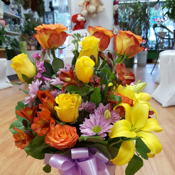 Mixed bouquet of yellow and orange roses with lilies and purple daisies in a glass vase