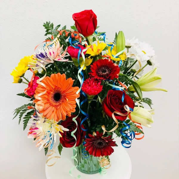 Colorful mixed bouquet with roses, gerbera daisies, lilies, and ribbon curls in a glass vase