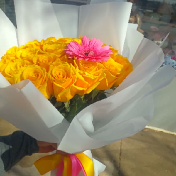 Handheld bouquet of yellow roses with a single pink gerbera wrapped in white paper with pink and yellow ribbons.