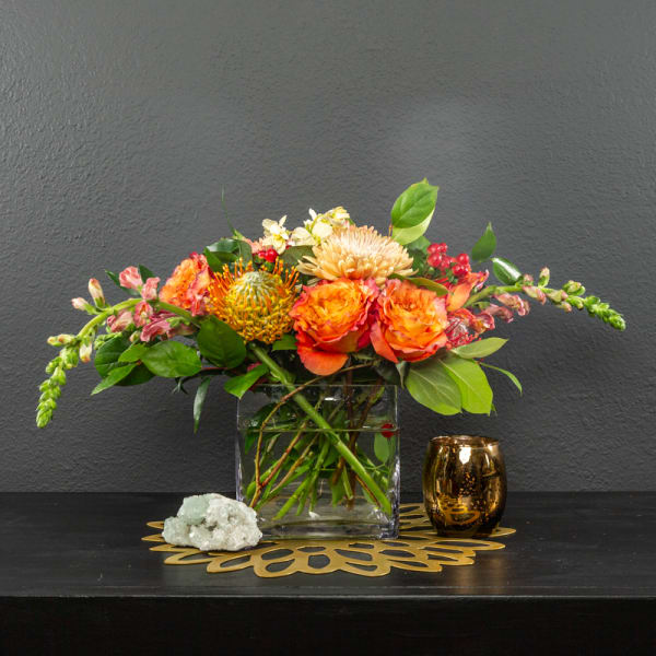 Orange and peach flowers arranged in a clear square vase with a gold candle holder.