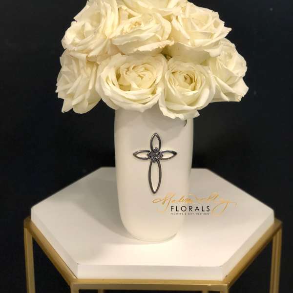 White roses arranged in a tall white vase with a cross emblem