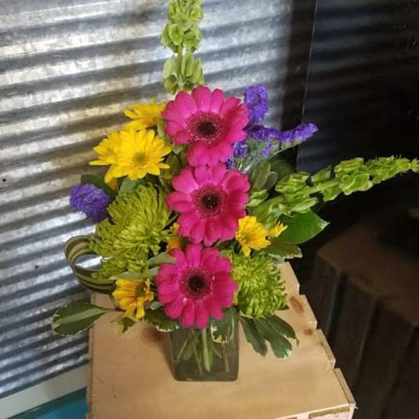 Bright mixed bouquet with pink gerberas, yellow daisies, and purple filler in a glass vase