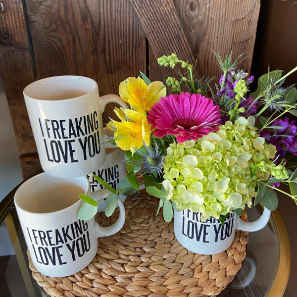 White 'I freaking love you' mugs, one filled with pink, yellow, and green flowers on a woven mat.