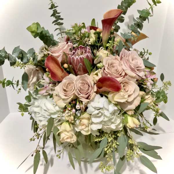 Round arrangement of blush roses, white hydrangeas, and orange calla lilies in a clear glass vase
