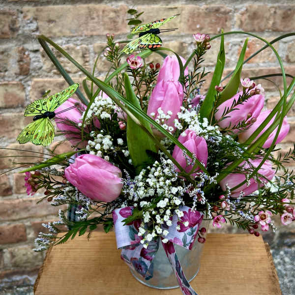 Pink tulip arrangement with white filler flowers, grass loops, and decorative butterflies in a metal pail.