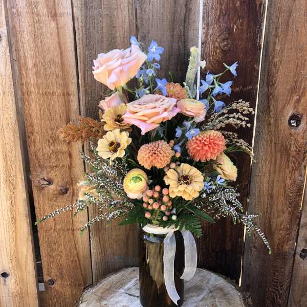 Rustic arrangement of peach roses, dahlias and mixed blooms in an amber glass jar with ribbon.