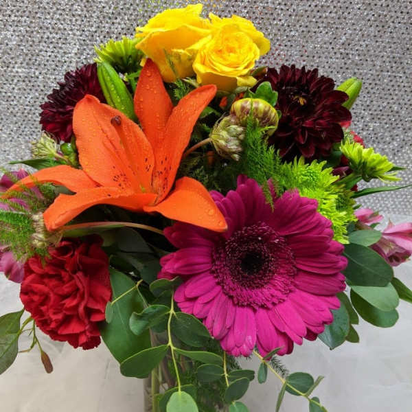 Colorful bouquet with orange lily, pink gerbera, yellow roses, and red carnations in a glass vase