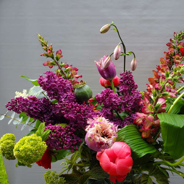 Tall mixed arrangement with purple lilacs, pink fringe tulips, and red blooms in a glass vase.