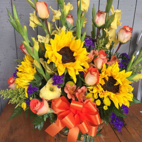 Tall basket arrangement with sunflowers, peach roses, yellow spikes, purple accents, and an orange bow.