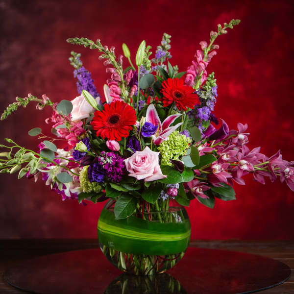 Mixed bouquet with red gerbera daisies, pink roses, and purple blooms in a green glass vase