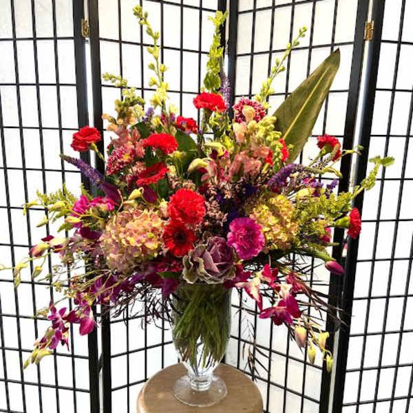 Large mixed flower arrangement in a clear glass vase on a stool