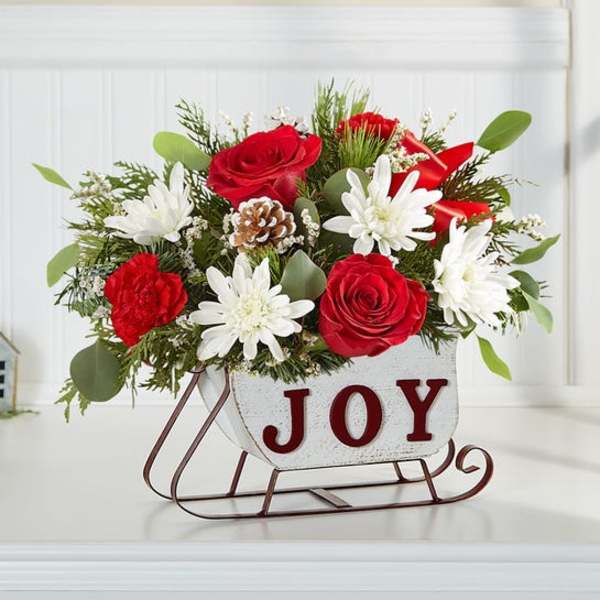 Red and white flower arrangement in a sleigh-shaped container with the word JOY