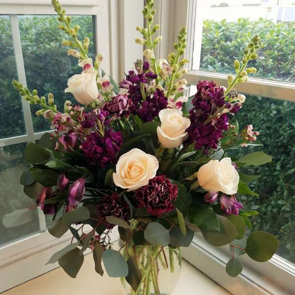 Tall arrangement of cream roses, purple flowers, and snapdragons in a clear glass vase