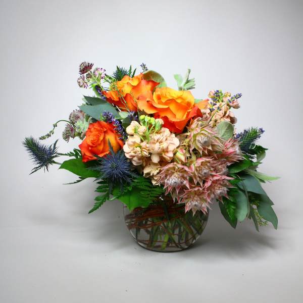 Orange roses arranged in a glass bowl with blue thistle and pale pink blooms
