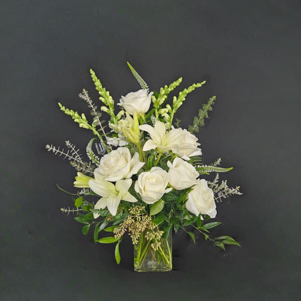 White roses, lilies, and snapdragons arranged in a clear glass vase against a dark background