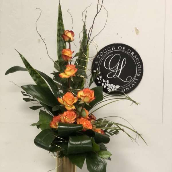 Tall orange rose arrangement in a gold vase with dark foliage