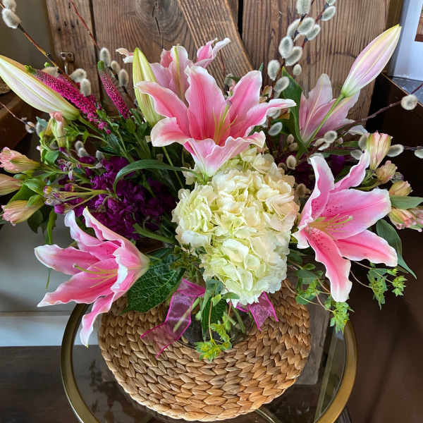 Arrangement of pink lilies, white hydrangea, and purple flowers in a vase with woven cover on a glass table.