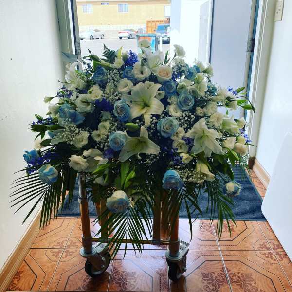 Large blue and white casket spray with roses and lilies displayed on a rolling cart indoors
