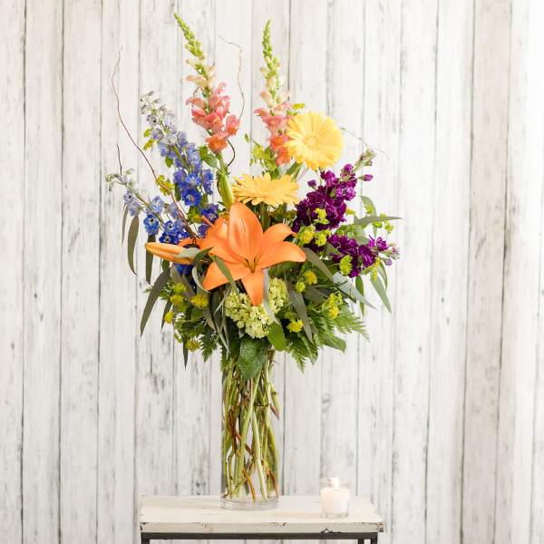 Tall mixed bouquet with orange lilies, yellow daisies, and purple blooms in a glass vase