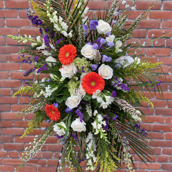 Standing spray of white roses, orange gerbera daisies, and purple blooms with tall palm fronds on an easel.