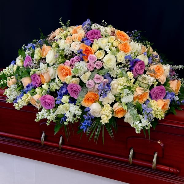 Large pastel casket spray of roses and mixed flowers in peach, purple, white, and blue on a wood casket