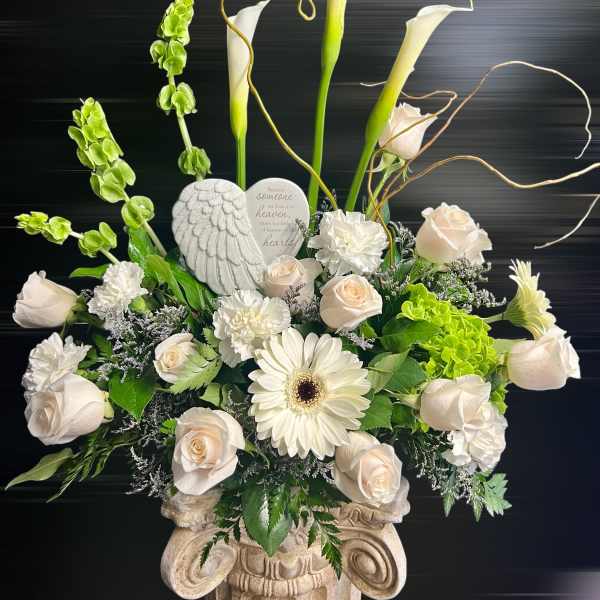 Tall white and green sympathy arrangement with roses, calla lilies, and angel plaque in a stone urn.