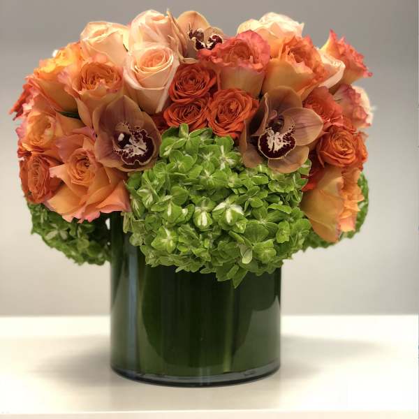 Compact arrangement of orange roses, orchids and green hydrangeas in a glass cylinder vase