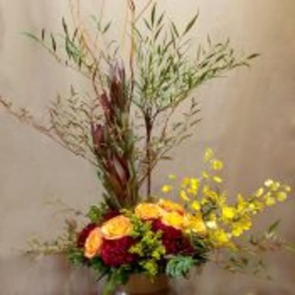 Tall autumn-toned arrangement with orange roses, red blooms, yellow flowers and branches in a brown vase