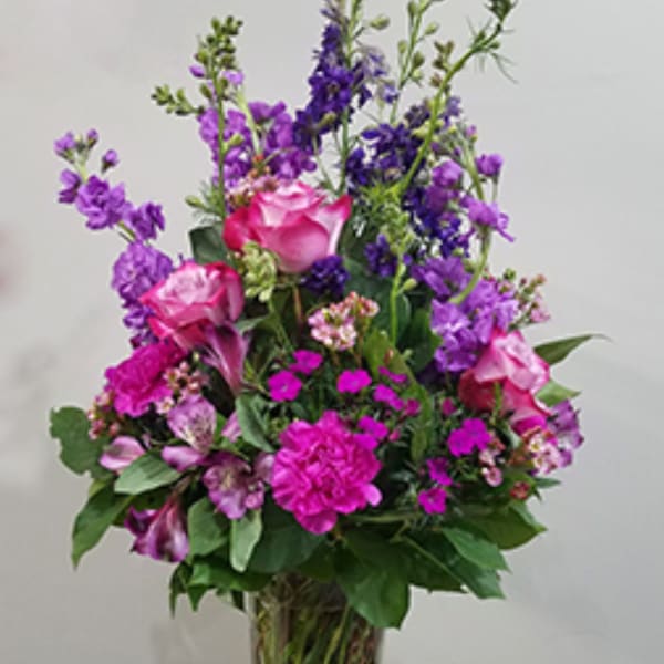 Tall arrangement of pink roses, bright pink carnations, and purple flowers in a clear glass vase