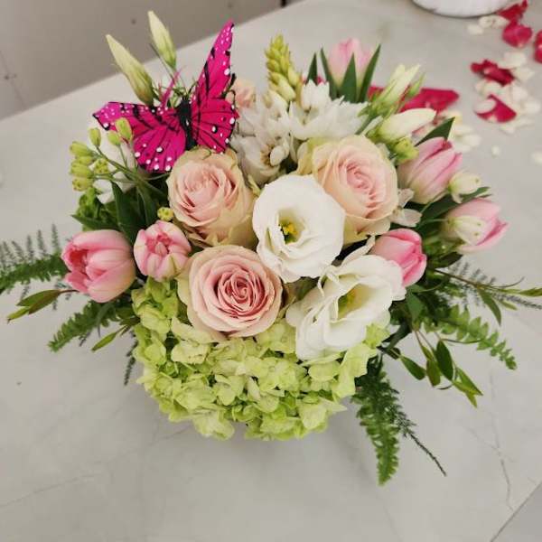 Round arrangement of blush roses, white blooms, pink tulips, and green hydrangeas with a pink butterfly pick