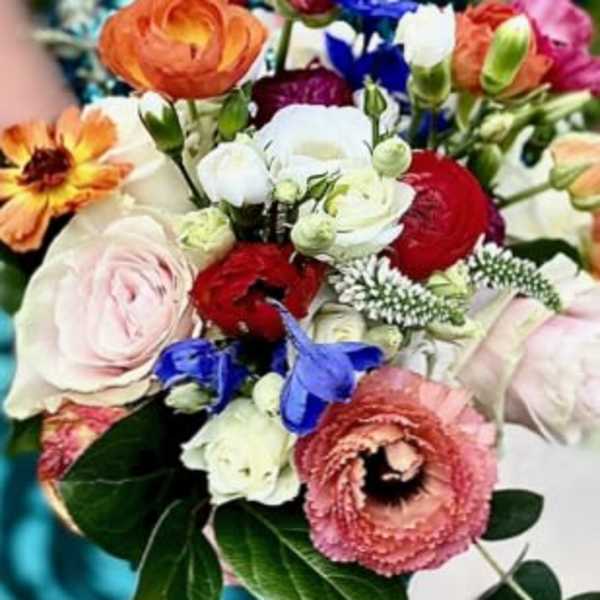 Colorful hand-tied bouquet with orange ranunculus, blush roses, red blooms, and blue accent flowers.