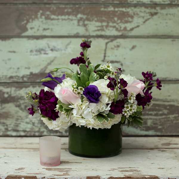 Purple and white floral arrangement in a dark green vase with a small candle nearby