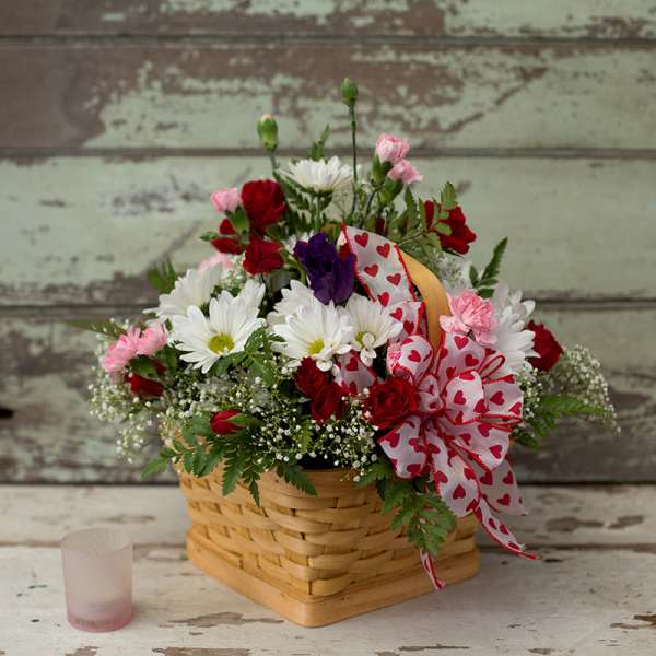 Basket arrangement of red roses, white daisies, and pink carnations with a heart ribbon