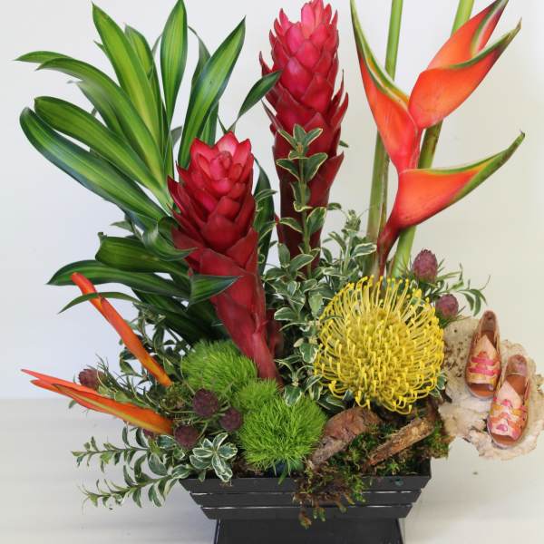 Tropical arrangement with red and orange blooms, yellow pincushion protea, and lush foliage in a black container