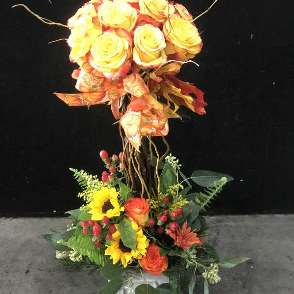 Tall floral arrangement with yellow and orange roses, sunflowers, and mixed greenery in a vase.