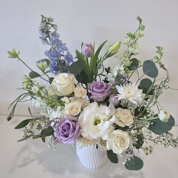 Pastel arrangement of white and lavender roses, lisianthus and delphinium in a white ribbed vase.