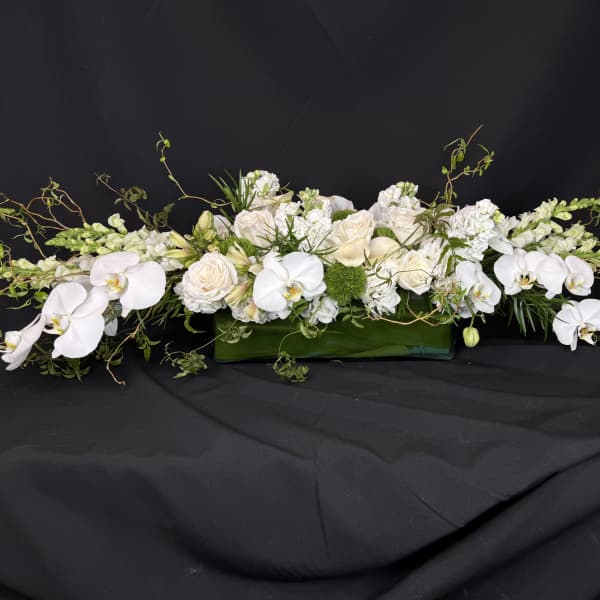 Long white floral arrangement with orchids and roses in a green rectangular container