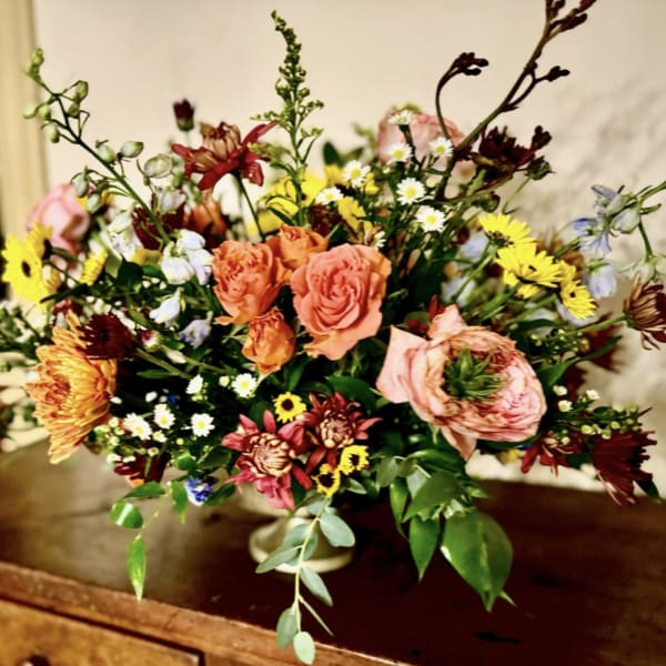 Low compote arrangement of peach roses, yellow daisies, and mixed flowers on a wooden table.