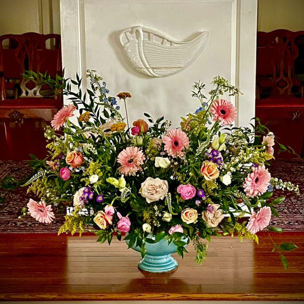 Large mixed pastel flower arrangement in a turquoise pedestal bowl before a white pulpit.