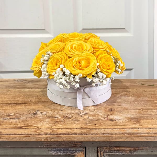 Round white box of yellow roses with small white filler flowers on a wooden table