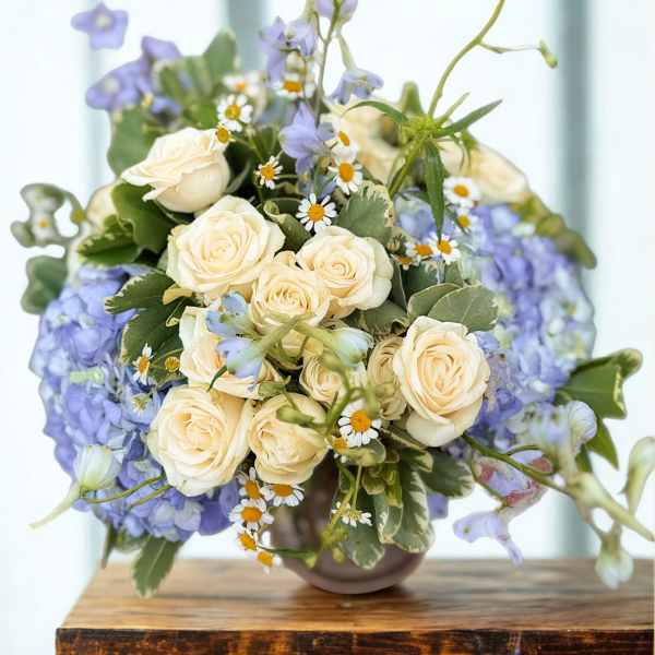Compact arrangement of cream spray roses, lavender hydrangeas, and small daisies in a round vase.