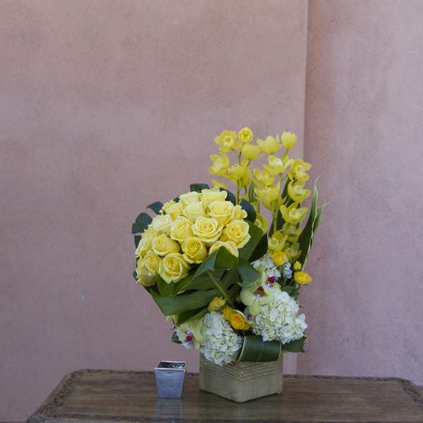 Yellow roses and orchids arranged in a square vase