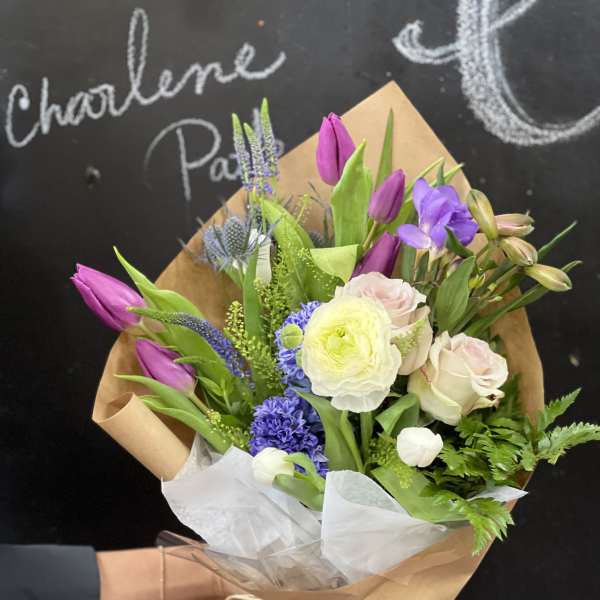 Hand-tied bouquet of purple tulips, pale roses, and mixed blooms in brown paper