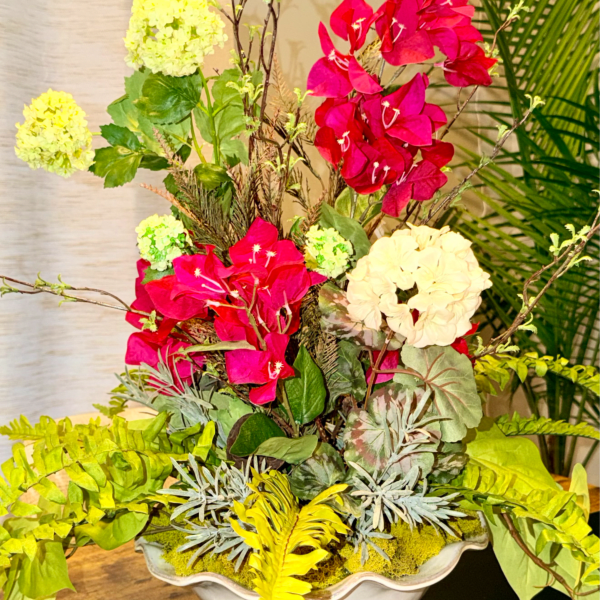 Tall arrangement of bright pink blooms, pale hydrangeas, and green ferns in a white ceramic bowl.