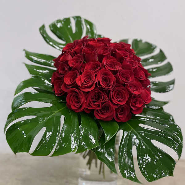 Compact dome of red roses framed by large glossy tropical leaves in a clear glass vase.