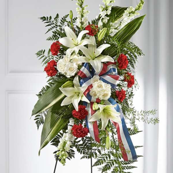 Tall standing spray with white lilies, white roses, red carnations and a red, white, and blue ribbon on an easel