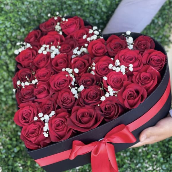 Heart-shaped black box filled with red roses and small white blooms, tied with a red ribbon.